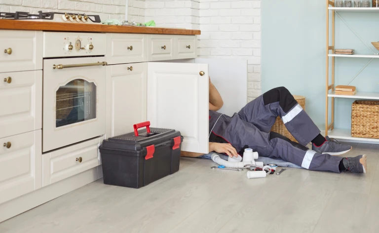 Plumber lying on the kitchen floor working under an open sink cabinet, with tools and plumbing parts scattered nearby and a toolbox beside the cabinets