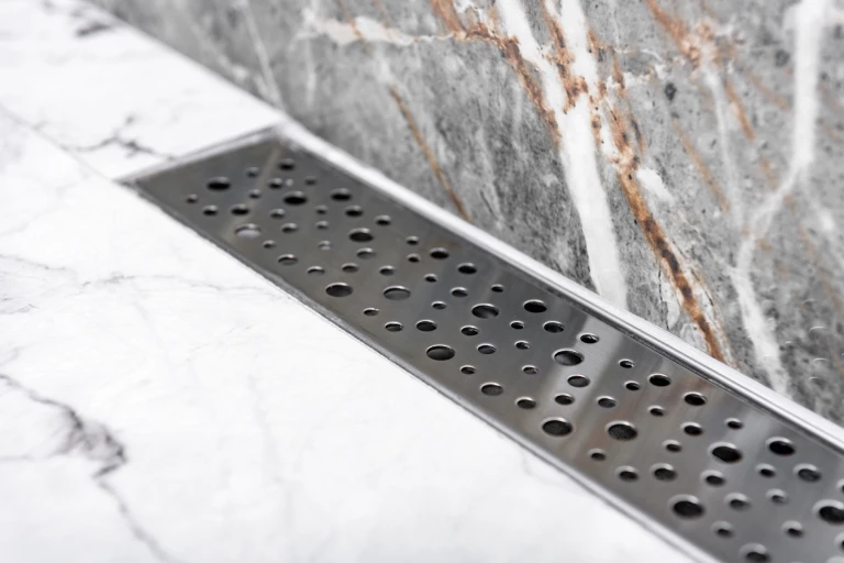 A sleek, rectangular stainless steel linear drain with a circular pattern installed along a white marble shower floor
