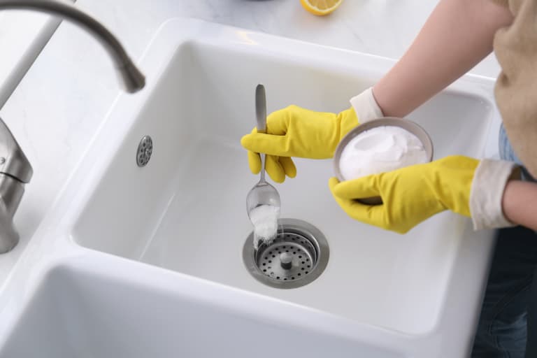 Yellow-gloved hands adding baking soda to a sink drain.