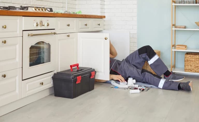 A repairman lying on a kitchen floor beneath an open sink cabinet.