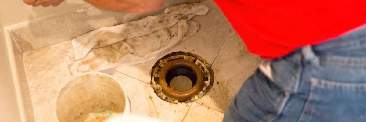 A plumber wearing a red shirt and jeans installing a new toilet flange
