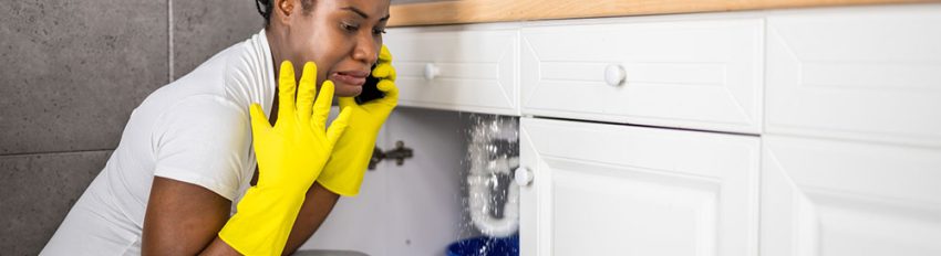 A woman on a call crouches in front of a sink with leaking pipes