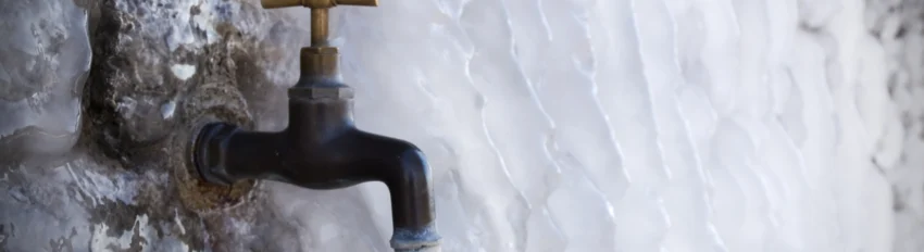 Outdoor faucet frozen and partially iced over in winter, with water dripping from the spout against an ice-covered wall